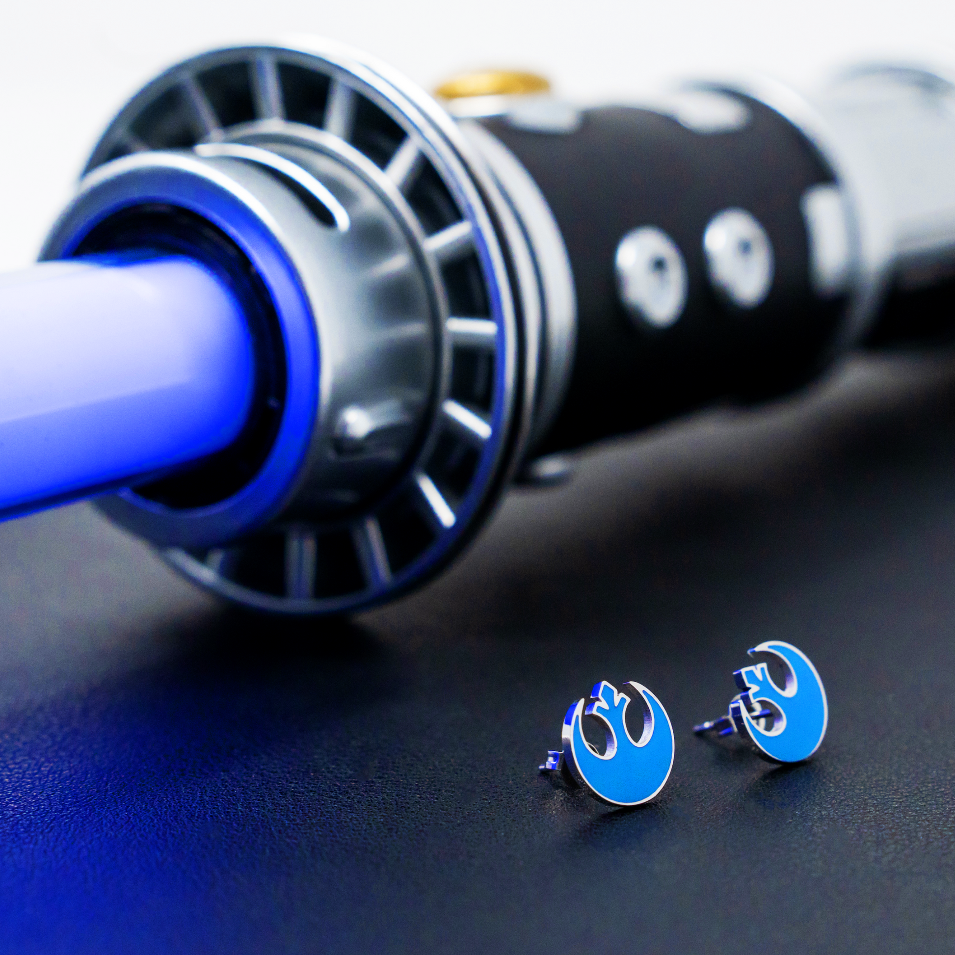 A pair of blue earrings featuring the rebel icon from Star Wars on a black backdrop with a blue lightsaber in the background
